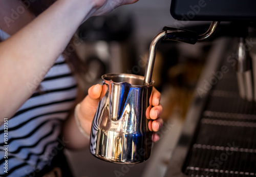 Barista steaming milk in a stainless steel pitcher using an espresso machine steam wand. Close-up view of coffee preparation process in café environment.