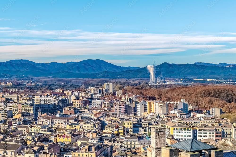 Fototapeta premium Panoramic winter view of Girona, Spain, with colorful buildings, autumn foliage, and the Pyrenees in the background. Steam from industry rises beyond the dense urban landscape