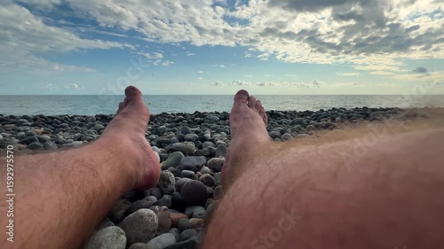 Horizontal 4K travel wide angle video close up. Male hairy bare legs shoes on Black Sea pebble beach. Sun day. Blue cloudy sky. Ocean waves. Concept of summer vacation, tourism, coast line, rest