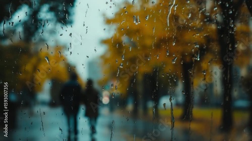 Two people walking in the rain, blurred view through a wet window. Yellow autumn leaves on trees. Urban setting with a rainy atmosphere.