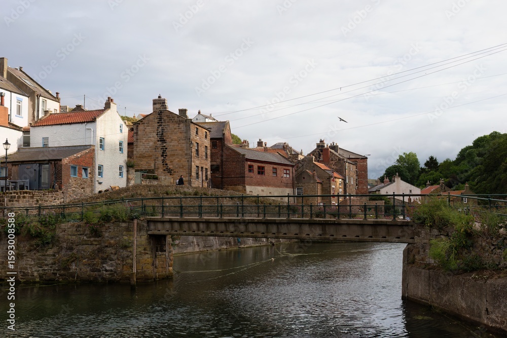 Fototapeta premium A picturesque view of a town in England. Buildings line a river, with a pedestrian bridge in the foreground in Staithes - North Yorkshire - United Kingdom
