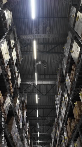 Upward view of a tall warehouse aisle filled with stacked pallets and cardboard boxes on metal shelving under industrial lighting and ventilation pipes.
