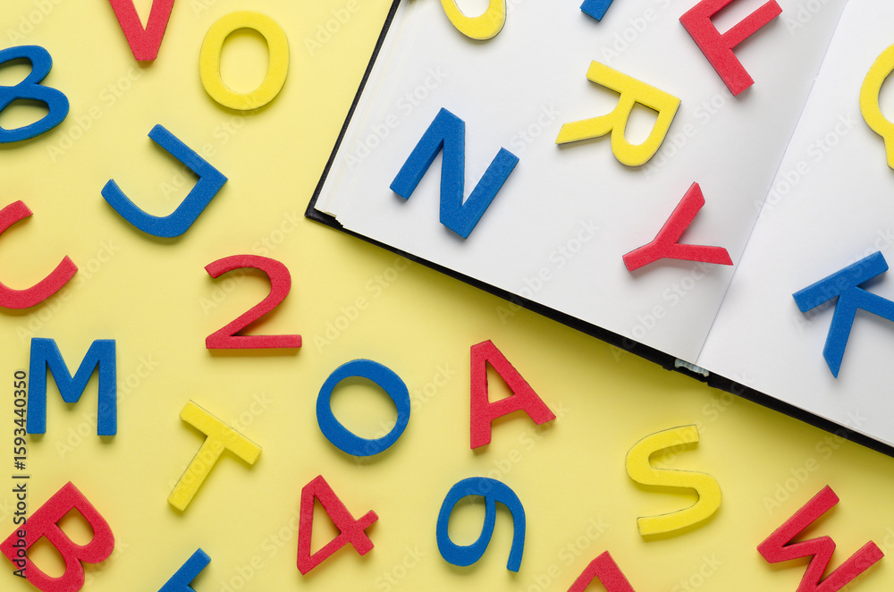 Fototapeta premium A notebook and colorful alphabet letters and numbers scattered on light yellow background. Back to school concept.