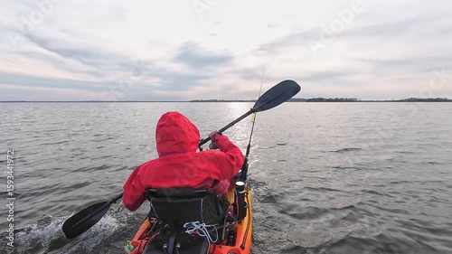 Fisherman sitting in a kayak and trolling to the secret spot to catch fish, while holding paddle. Kayak fishing competition. Angler in dry suit or drysuit for environmental protection and cold water.