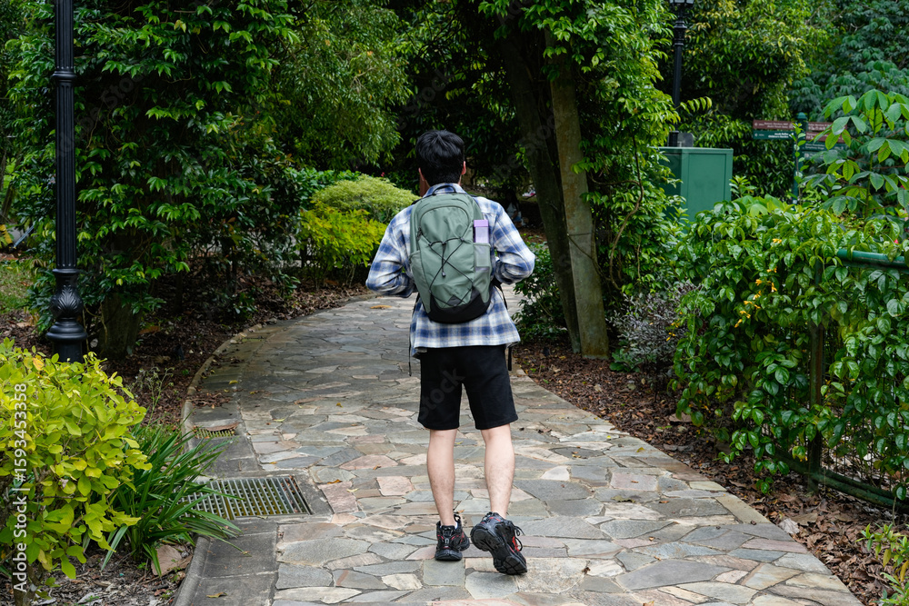 Fototapeta premium Young man with a backpack walking on a winding path in a lush, green park or garden. Focus on nature, outdoor activity, and solo exploration.