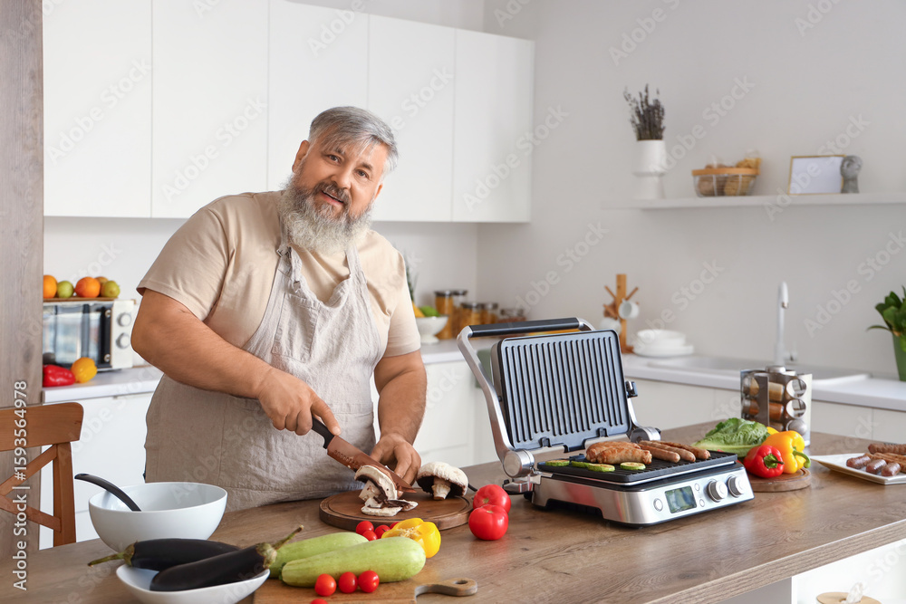 Mature man cooking tasty sausages on modern electric grill and cutting fresh vegetables at table in kitchen Mature man cooking tasty sausages on modern electric grill and cutting fresh vegetables at table in kitchen
