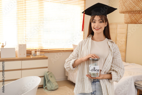 Young woman in graduation cap holding college savings jar with money, educati...