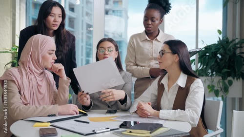 Serious multiethnic women females work team girls African American Japanese Caucasian Arabian Muslim hijab businesswomen analyze document graph low data brainstorm strategy inside office meeting room