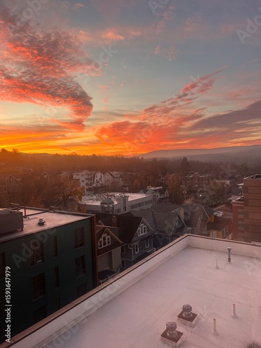 Spectacular fiery sunset with dramatic orange and pink clouds over small town rooftops viewed from white rooftop terrace creating breathtaking urban evening sky