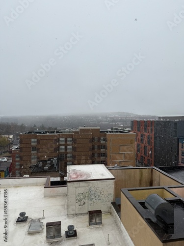 Urban winter snowfall scene showing heavy snow falling over city rooftops and apartment buildings on an overcast gray day, captured from elevated perspective with HVAC equipment and residential archit