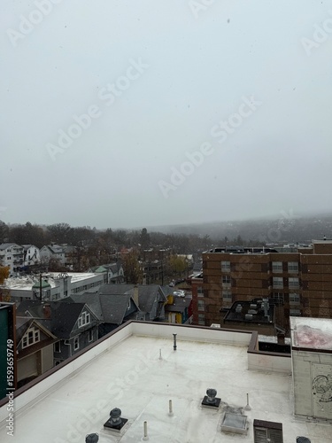 Panoramic winter cityscape showing mixed residential neighborhood with Victorian houses, modern apartments, and snow-covered rooftops during overcast weather, viewed from flat rooftop perspective with