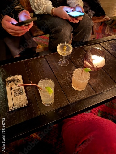 Friends enjoying cocktails and socializing while using smartphones at rustic wooden bar table with craft drinks, lime garnishes, and QR code menu in cozy restaurant atmosphere