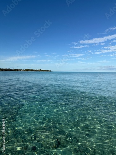 Crystal clear tropical ocean water with visible sandy bottom creating mesmerizing light patterns, distant palm-lined coastline under expansive blue sky with scattered white clouds in Caribbean