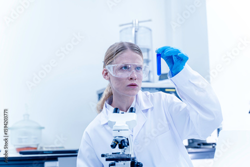 Young female scientist holding test tube filled with blue liquid, carefully reviewing experiment data related to science, laboratory, scientist, experiment, and research.