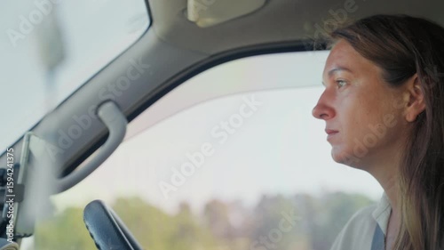 Close-up of a young adult woman focused on driving a car under clear blue skies with sunlight filtering through the trees outside.