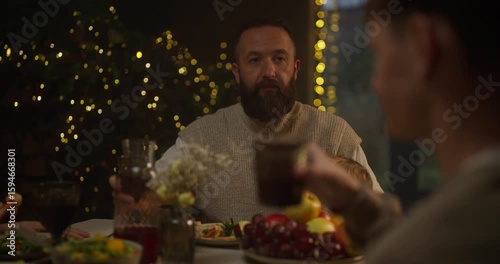 A brunette man with a beard holds his little daughter in his arms and clinks a glass with his son at the Christmas table at home