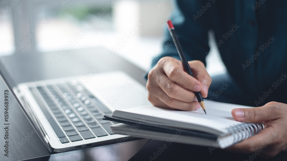 Fototapeta premium Student studying online class via laptop computer and writing note on paper notebook. Business woman working on laptop computer, searching the ideas and taking note, e-learning, close up