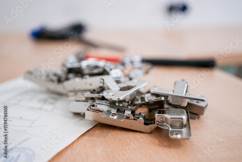 Many furniture hinges are lying on top of a kitchen project on a wooden worktop, ready for use in assembling new furniture, with some tools visible in the background out of focus