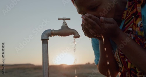 Backlit cropped close-up of a Black African woman in traditional clothing drinking water from a faucet at sunset