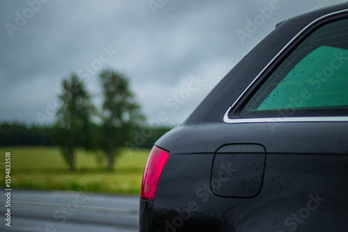 Canvas-taulu Close-up of a wet black car's rear with taillight and window, parked beside countryside road under overcast sky