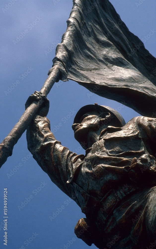 Fototapeta premium A monument of an Asian soldier holding up the flag, against a blue sky background, shot from a low angle with a wide-angle lens, in a sculptural style with bronze material, displaying a strong contras