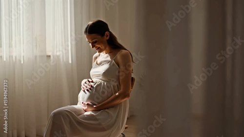 A pregnant woman in a flowing white dress cradles her belly, standing serenely in soft sunlight