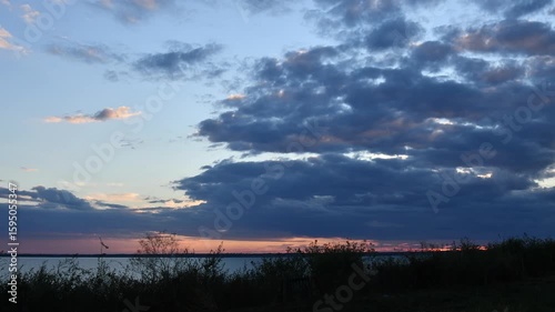 Transition of day to night as clouds drift across expansive sky during sunset over serene lake water. Colors shift dramatically, illuminating the horizon.