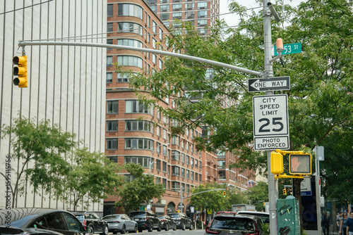 A City Speed Limit 25 sign with a photo enforcement notice stands near a traffic light on a busy urban corner. Modern buildings, cars, and a street sign for 53rd Street are visible in the background.
