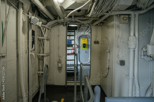 Foto A narrow corridor inside a military ship shows exposed cables, metal pipes, and a yellow safety sign on a door in the background