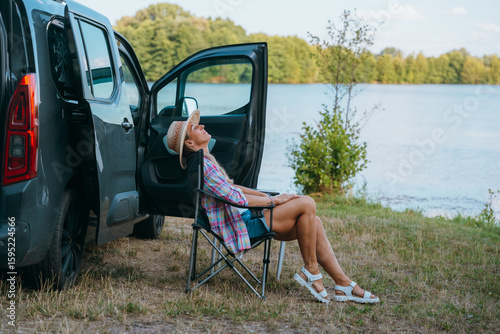 Fotografie Woman sits peacefully beside her campervan at a quiet lakeside spot, surrounded by nature