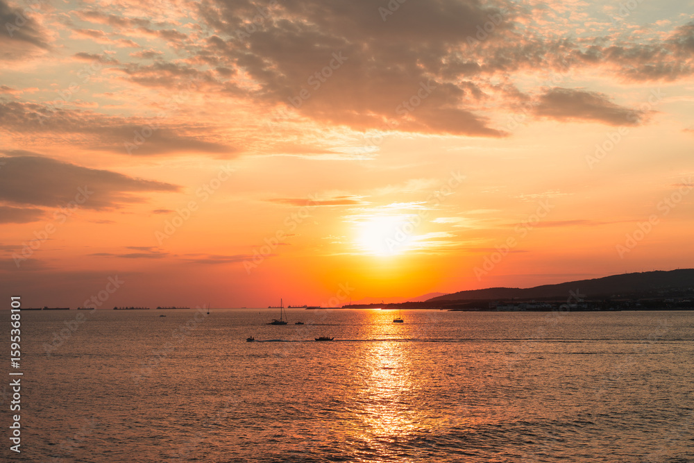 Fototapeta premium Beautiful sunset over the sea with golden sunlight reflecting on the water, small boats on the horizon, and distant coastline under a dramatic sky.