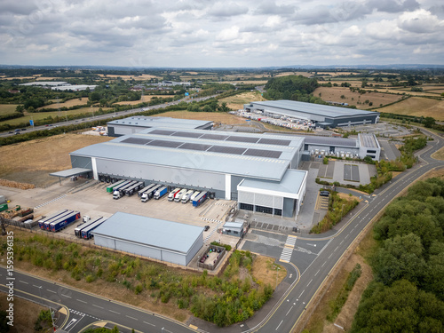 Large UK Logistics and Distribution Centre with Delivery Trucks, Solar Panels and Parking, Captured by Drone in Rural Industrial Estate Alongside Main Road Infrastructure and Surrounding Countryside
