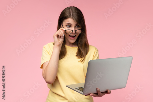 Studio shot of a happy young casually dressed woman holding using laptop computer and looking at its screen with excited face expression, isolated over pastel pink background