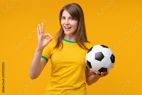 Young woman on yellow studio background holding a soccer ball and showing an OK hand gesture, expressing confidence, positivity, and support as a cheerful fan or athlete