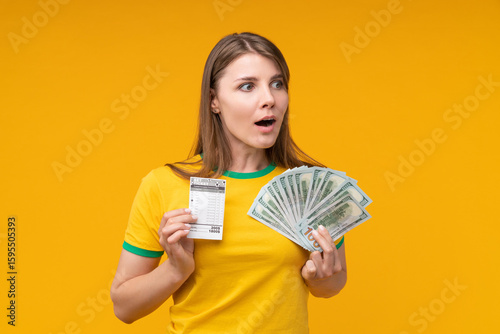 фотографія Astonished young woman posing on yellow background, holding a betting ticket in