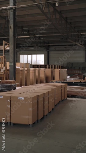 Cardboard boxes arranged in rows inside industrial warehouse. Vertical video of an indoor warehouse showing rows of neatly arranged cardboard boxes, surrounded by wooden furniture parts and industrial