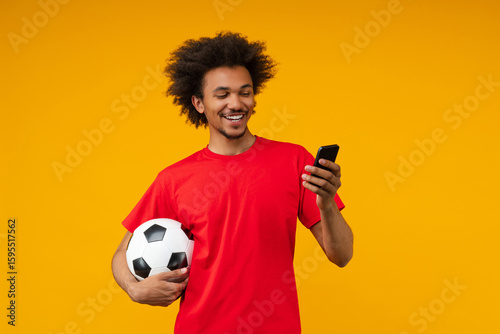 Smiling young African man holding a soccer ball and looking at his smartphone screen