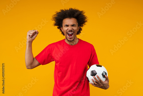 Excited African man on yellow background screaming with rage and passion while holding soccer ball and making a yes gesture