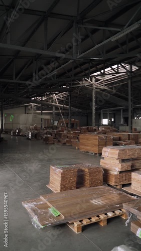 Wide shot of a spacious industrial warehouse storing large stacks of wooden boards and pallets. The camera moves slowly through the clean, well-lit interior with steel trusses and a concrete floor. 