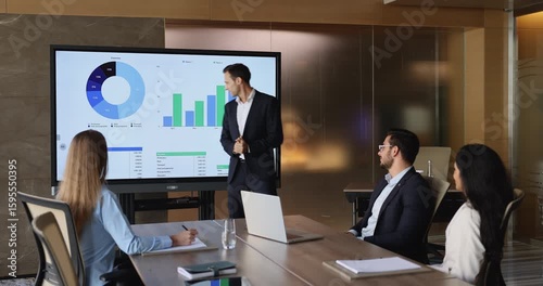 Confident project manager man presenting marketing sales report on large presentation screen, pointing at analytic chart, talking to colleagues sitting at meeting table