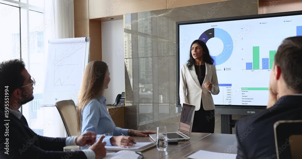 Young Indian business product owner woman discussing analytics, marketing research result with team, presenting sales financial infographics on large wall TV screen, talking to colleagues