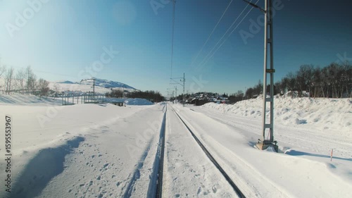 A scenic view of a snow-covered railway cutting through a winter landscape, with overhead electric lines and distant mountains under a clear blue sky. Peaceful and cold rural setting.