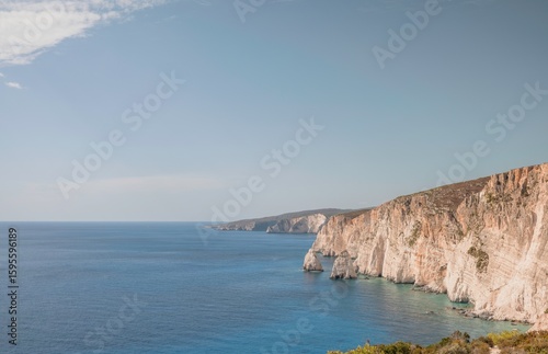 Fototapeta Naklejka Na Ścianę i Meble -  Under a pale blue sky, the turquoise sea kisses the rugged cliffs of Zakynthos. Sunlight paints the towering bluffs in warm tones as waves crash gently against the shore