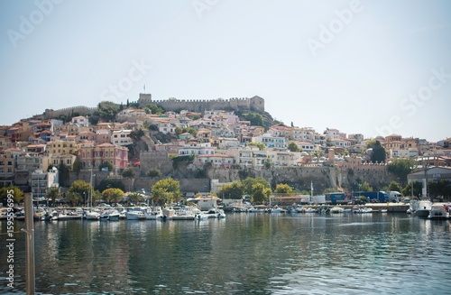 On a bright day in Kavala, Greece, colorful buildings cascade down a hillside, culminating in a marina full of boats. An old fortress looms over all