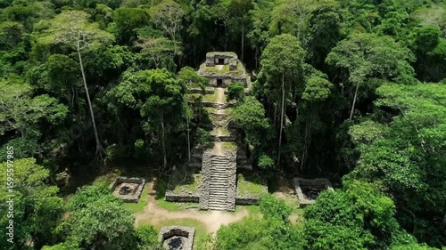 An ancient stone temple with long steps rises majestically from the dense, lush green jungle canopy, viewed from an aerial perspective under a partly cloudy sky
