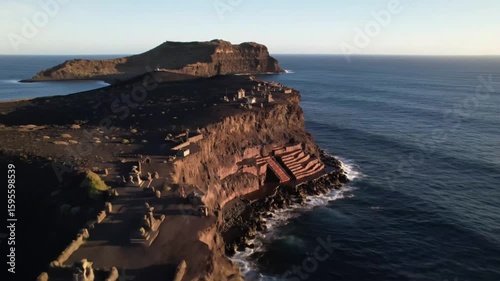 Dramatic coastal cliffs meet the vast blue ocean under a clear sky at golden hour, highlighting ancient structures