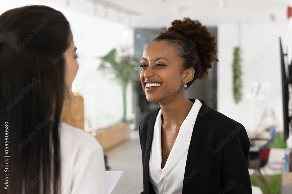 Obraz premium Happy African business woman sharing joyful or humorous moment with female colleague, laughing at joke or personal story, enjoy lighthearted and friendly conversation during break in coworking space