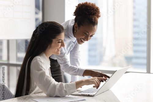 Two smiling multiracial women colleagues pointing at laptop screen together, laughing while reviewing amusing content, enjoy teamwork successful result during informal meeting or brainstorming session