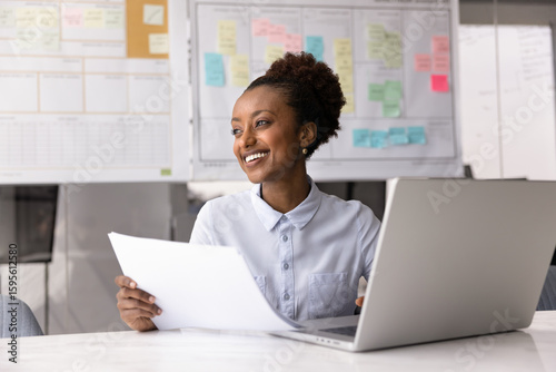African woman employee sits at desk holding documents distracted from laptop work, look into distance with contented smile, review positive feedback or results, feel satisfied with professional growth