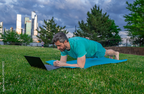Online fitness. Online workout on yoga mat outdoor. Mature man training with online fitness video. Man watching exercise tutorial on laptop. Man practicing plank in city park. Online fitness lesson.